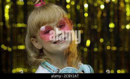 Child dances, listens music. Relaxing, enjoying, having hun. Little kid girl 5 years old in sunglasses and with headphones isolated on golden shiny background in studio Stock Photo