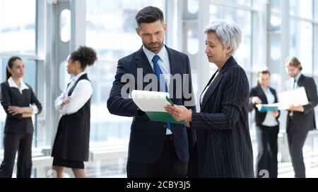 Businessman and businesswoman discussing work in office hall with colleagues on background. Stock Photo