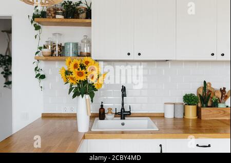 View on clean white simple modern kitchen in scandinavian style, kitchen details, wooden table, sunflowers bouquet in vase on the table Stock Photo