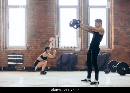 young man doing exercises with kettlebell while his female friend performing squats. training process in the sport center.spare time Stock Photo