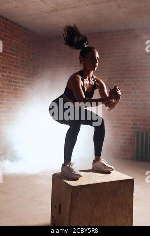 Attractive sporty female with long hair doing squats on wooden box. Process of jumping. Brickwall background Stock Photo
