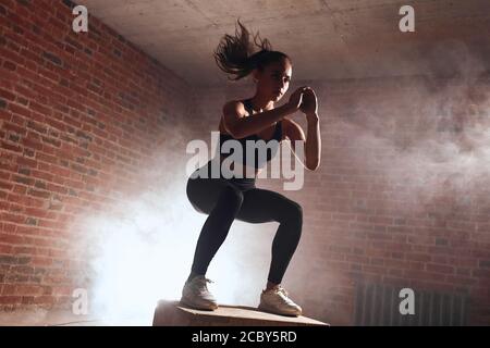 Attractive sporty female with long hair doing squats on wooden box. Process of jumping. Brickwall background Stock Photo