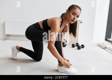 tying shoelashes in gym. Flexible girl in black leggins and topic preparing to do exercises and look at camera Stock Photo