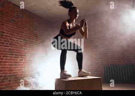Attractive sporty female with long hair doing squats on wooden box. Process of jumping. Brickwall background Stock Photo