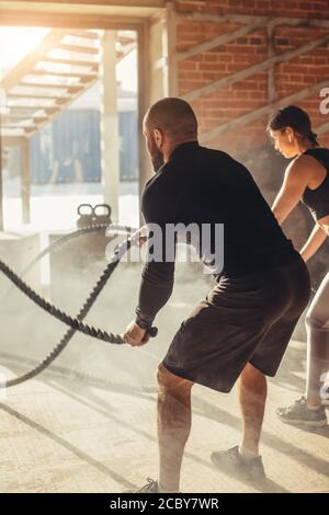 Caucasian fit attractive couple in sports clothing concentrated on the exercise with battle rope requiring increased effort and strength, standing at Stock Photo
