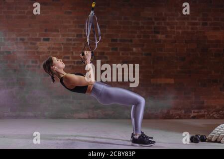 Young attractive woman training with trx fitness straps in the crossfit studio, suspension systems of ropes or belts that allow sportgirl to perform e Stock Photo