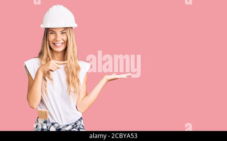 Beautiful caucasian woman with blonde hair wearing hardhat and painter clothes amazed and smiling to the camera while presenting with hand and pointin Stock Photo