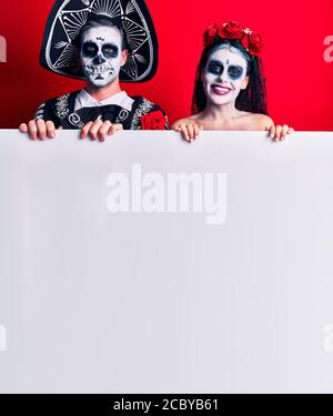 Young couple wearing mexican day of the dead costume holding blank empty banner looking positive and happy standing and smiling with a confident smile Stock Photo