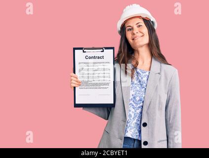 Young beautiful brunette woman wearing architect hardhat holding contract looking positive and happy standing and smiling with a confident smile showi Stock Photo