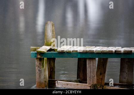 A worn and textured wooden plank wharf. The boards are grey and one horizontal green one with smooth water in the background. Stock Photo