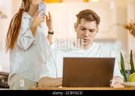 young caucasian woman talk on phone while her husband work in laptop, freelance work. married couple busy with their own business, quarantine during c Stock Photo