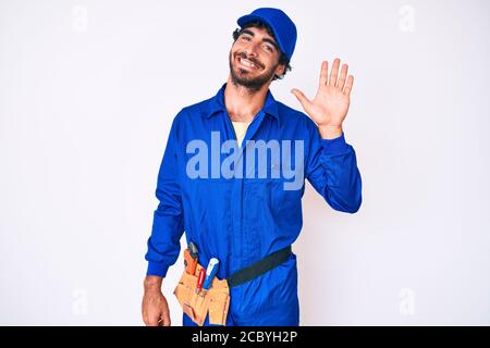 Handsome young man with curly hair and bear weaing handyman uniform waiving saying hello happy and smiling, friendly welcome gesture Stock Photo