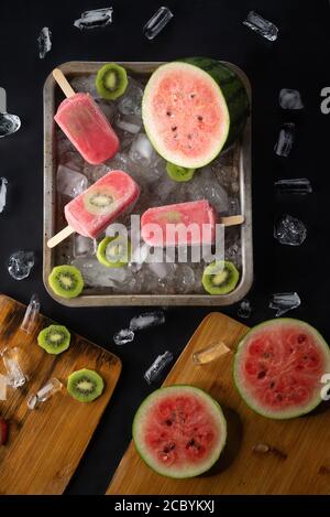 Homemade watermelon and kiwi ice cream or popsicles garnishes with fruits and pieces of ice on black table Stock Photo