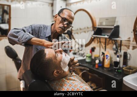 MARRAKESH, MOROCCO - SEPTEMBER 10, 2019: Man shaving in traditional street arabian barbershop in Marrakech. Local barber job in Morocco. Stock Photo