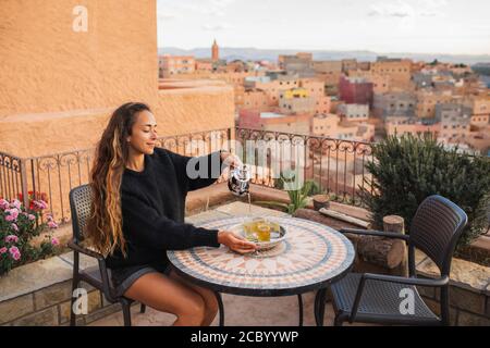 Woman pouring traditional moroccan mint tea on terrace with amazing view of old arab town Boumalne in Morocco at sunset. Round vintage table. Stock Photo