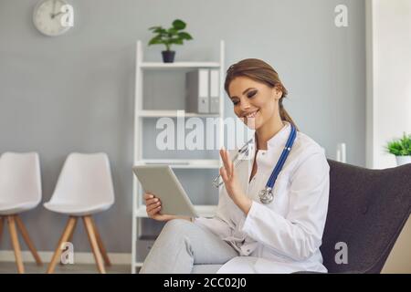 Doctor waving hand has video chat with patient using tablet in clinic office. Stock Photo