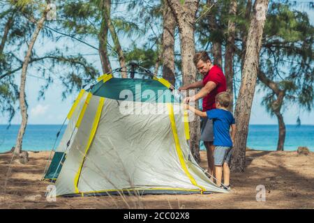 Camping people outdoor lifestyle tourists put up set up green grey campsite summer forest near lazur sea. Boy son helps father study mechanism of Stock Photo