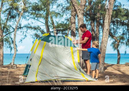 Camping people outdoor lifestyle tourists put up set up green grey campsite summer forest near lazur sea. Boy son helps father study mechanism of Stock Photo