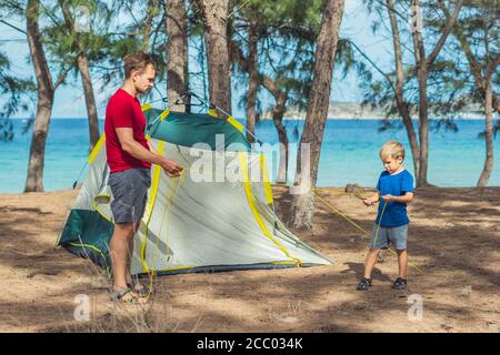 Camping people outdoor lifestyle tourists put up set up green grey campsite summer forest near lazur sea. Boy son helps father study mechanism of Stock Photo