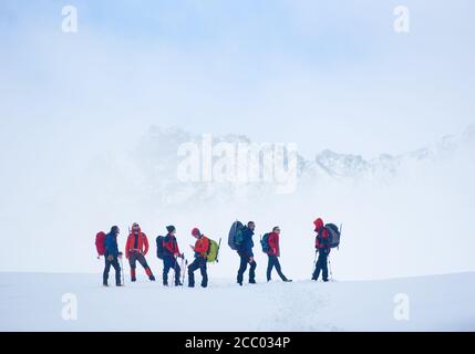 Hikers team with backpacks standing on hillside valley covered with snow, admiring the view of winter mountains while taking rest break during hike. Concept of travelling, hiking and mountaineering. Stock Photo