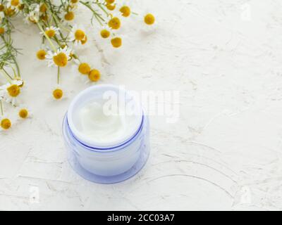 Cosmetic cream in a glass can and fresh chamomile flowers on the white structured background. Top view. Stock Photo