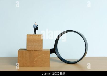 Miniature people concept - a desperate businessman standing on wood block thinking for solution with magnifier glass Stock Photo