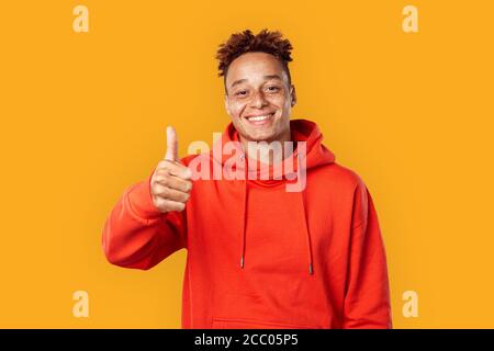 Freestyle. Mulatto freckled guy in hoodie standing isolated on yellow showing thumb up smiling cheerful Stock Photo