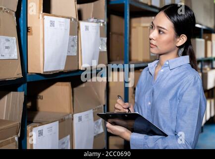 Close up of Asian smart businesswoman working with product inventory and writing on clipboard in distribution warehouse. For business warehouse invent Stock Photo
