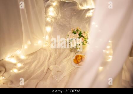 Soft plaids on the grass, bouquet with roses, plate with croissants, white canopy on the Tree. Evening photo zone in nature. Stock Photo