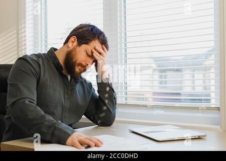 Tired thinking economist working with documents at office, closed laptop on desk. Stock Photo