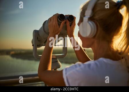 Woman in headphones using coin operated binoculars on the street Stock Photo