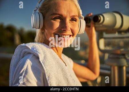 Cheerful woman in headphones using observation binoculars on the street Stock Photo