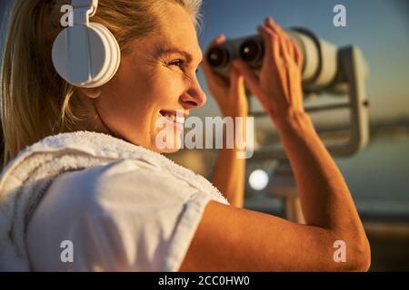 Joyful woman in headphones using observation binoculars on the street Stock Photo
