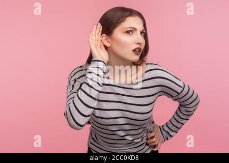 What? I can't hear you! Portrait of confused woman in striped sweatshirt keeping hand near ear to listen better, having hearing problems, difficult to Stock Photo