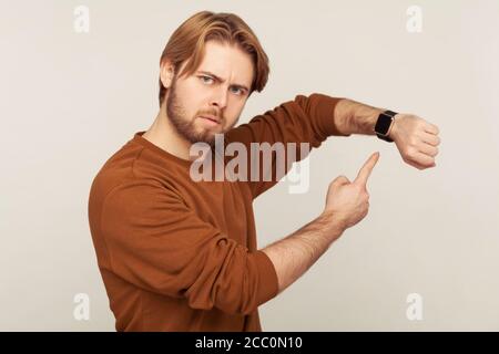 Look at time! Portrait of angry impatient man with beard in sweatshirt pointing wrist watch and looking annoyed displeased, showing clock to hurry up. Stock Photo