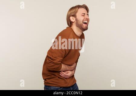 Hilarious laughter. Portrait of amused man with beard wearing sweatshirt holding belly and laughing out loud after hearing crazy anecdote, funny joke. Stock Photo