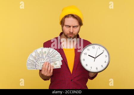 Angry hipster bearded guy in checkered shirt holding big clock and dollars banknotes, looking at camera with aggressive evil expression, frustrated ab Stock Photo