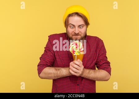 Hungry hipster guy in beanie hat and checkered shirt looking at lollipop with desire, struggling temptation to eat sweet sugary confectionery, delicio Stock Photo