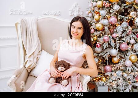 Woman in pink dress with small bear sitting on a chair near light Christmas tree. New Year, Christmas, Holiday Stock Photo