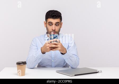 Amazed businessman sitting in office workplace reading shocking message on cellphone, using mobile application with surprised astonished expression. i Stock Photo