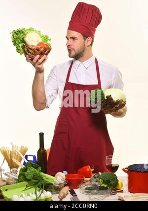 Chef in burgundy uniform holds baskets with cabbage and lettuce. Kitchenware and cooking concept. Cook stands by table with kitchenware and ingredients. Man with beard isolated on white background. Stock Photo