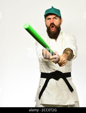 Man with beard in white kimono and green cap on white background. Violence and bullying concept. Hooligan gets ready to fight. Baseball player with angry face holds green baseball bat. Stock Photo