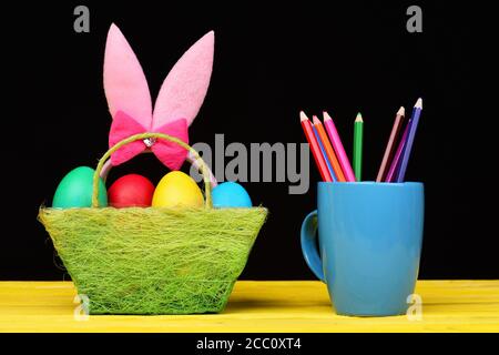 Girls Easter bunny ears in pink colour standing in green cloth basket filled with painted eggs and coloured crayons in blue mug placed nearby on wooden yellow table ,on black background. Stock Photo