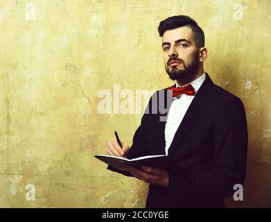 Bearded employer with cunning face expression and neat stylish outfit writes notes in his planner, cracked vintage beige background with copy space. Concept of planning and time management Stock Photo