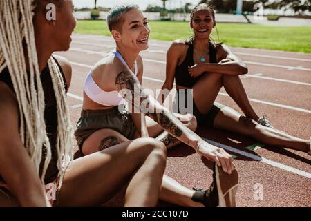 Group of three female runners taking a break from running workout and stretching their legs on race track. Athletes relaxing after running workout at Stock Photo