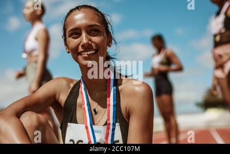 Female athlete with medal sitting on racetrack with team standing in background. Sportswoman looking away while sitting on athletics race track in sta Stock Photo