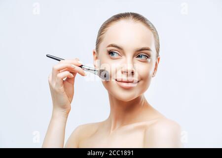 Facial treatment, cosmetology concept. Pretty young woman with dark combed hair, applies powder foundation on face with beauty brush, stands sideways, Stock Photo