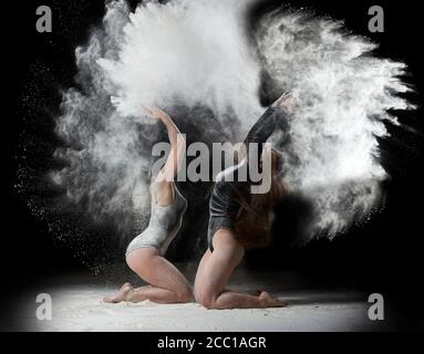 two beautiful young caucasian women in black bodysuits with a sports figure are dancing in a white cloud of flour on a black background, explosion and Stock Photo