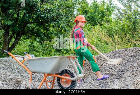 kid working on construction site. teen girl takes out rubble from wheelbarrow. kid with shovel loading crushed stones. Laying the foundation, building project. girl with wheelbarrow of rubble. Stock Photo