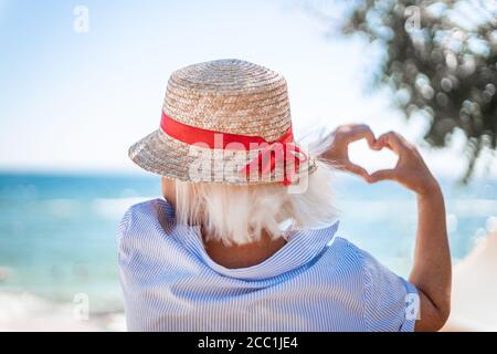 Blonde girl in a straw hat with her back makes love sign with her hands on blue sea background Stock Photo